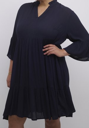 Woman wearing a loose, navy blue, tiered dress with three-quarter bell sleeves and a V-neckline, posing with one hand on her hip.