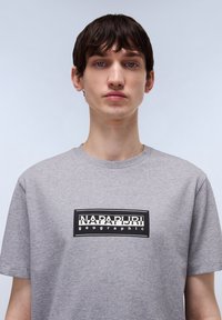 Camiseta de algodón gris con un gráfico rectangular negro en el pecho que dice "NAPAPIJRI geographic." Manga corta y cuello redondo.