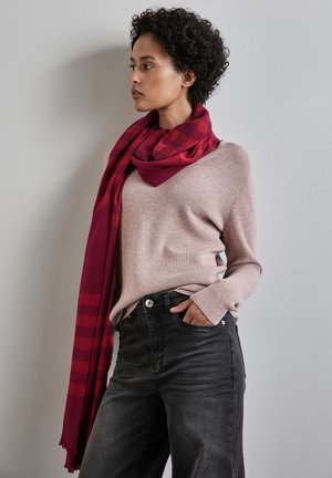 Een rood geruit sjaal gedrapeerd om een lichtroze ribbeltrui, gecombineerd met zwarte denim jeans. De outfit heeft een ontspannen pasvorm en gestructureerde stoffen.