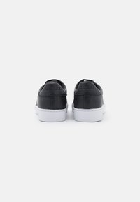Baskets en cuir noir avec semelles en caoutchouc blanc, dotées d'un design bas et d'un logo embossé sur le talon. Texture lisse, style minimaliste.
