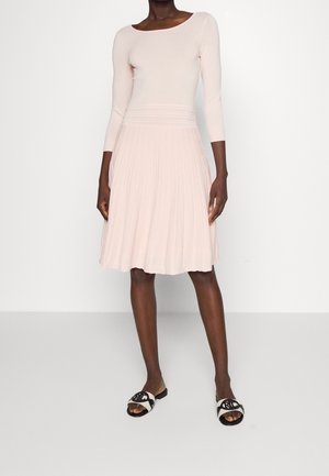 Woman wearing a pale pink fitted top and pleated skirt, paired with black and white slide sandals, standing against a plain white background.