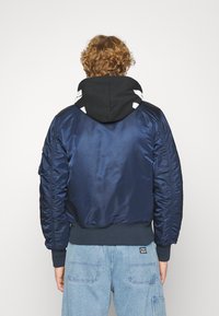 Tamno plava bombarder jakna s sjajnom teksturom, rebrastim manžetama i rubom. Ima crnu kapuljaču s bijelim detaljima i džep na lijevom rukavu.