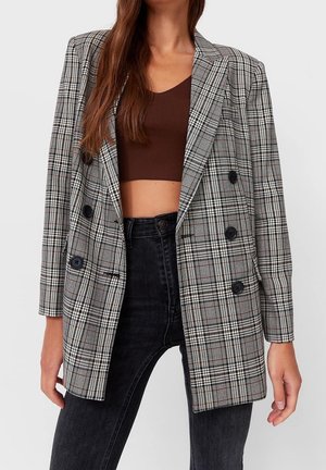 Kobieta ubrana w oversize'ową, szarą kraciastą dwurzędową marynarkę noszoną na krótkim brązowym topie i wysokich czarnych dżinsach, stojąca na jednolitym tle.