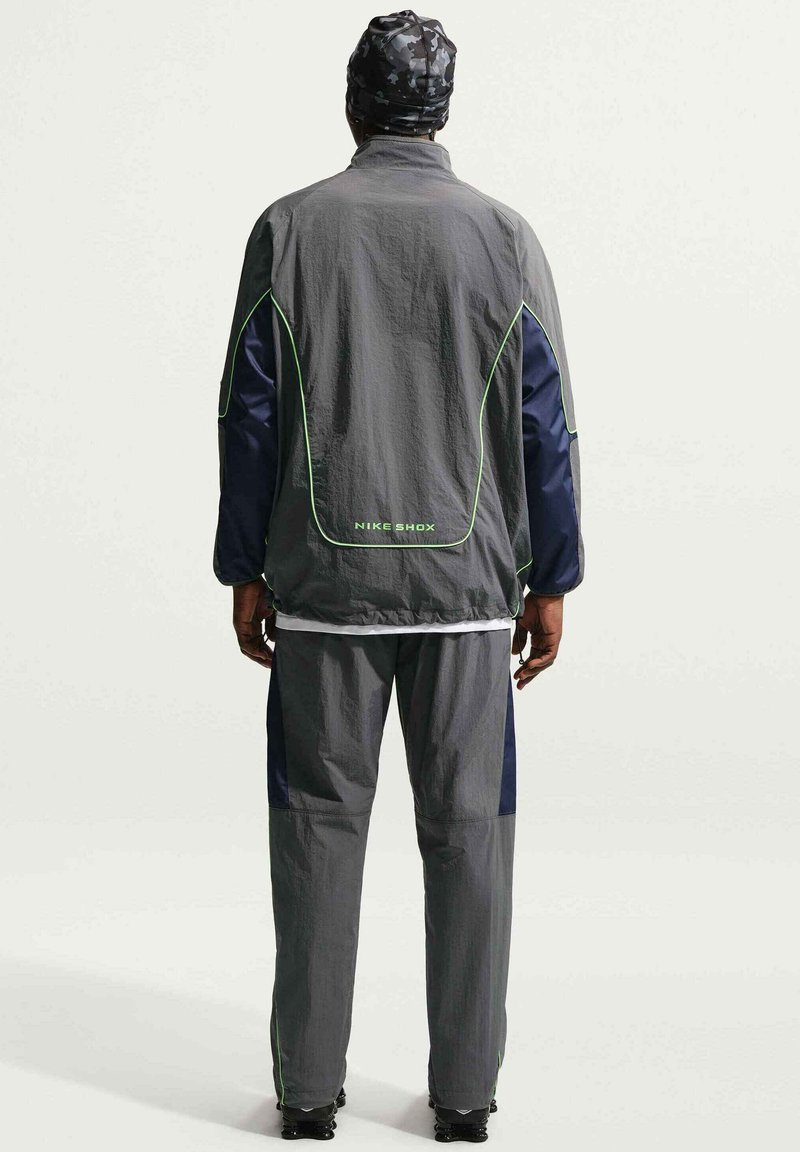 Persoon staat met de rug naar de camera, draagt een grijs trainingspak met groene bies en "NIKE SHOX" tekst, en een zwarte patroon hoofdbedekking.