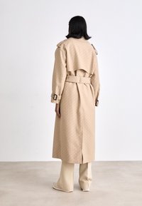 Manteau trench beige avec une texture à motifs, taille ceinturée et épaulettes. Manches longues et fente dans le dos pour une meilleure liberté de mouvement.