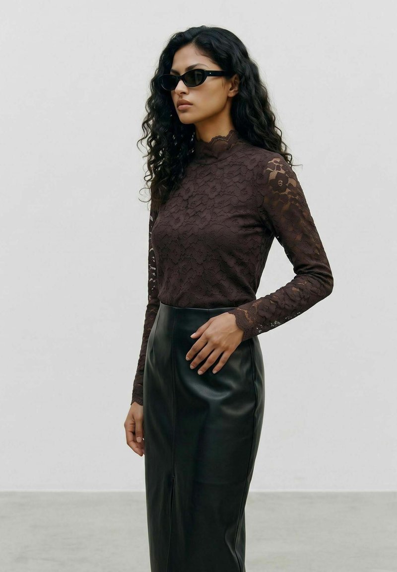 Femme aux longs cheveux bouclés portant des lunettes de soleil noires, un haut en dentelle sombre à manches longues et une jupe en cuir noire taille haute, debout de profil.