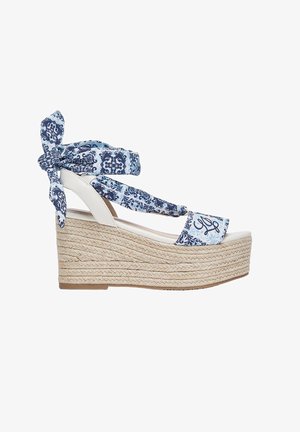 Sandaal met sleehak en gevlochten jute plateau, witte zool en blauw gemêleerde stoffen bandjes vastgeknoopt rond enkel en voet.