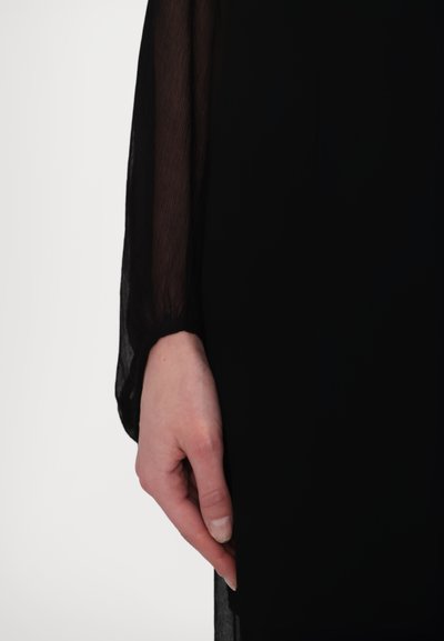 Sort kjole med gennemsigtige, lange ærmer. Stoffet ser let og let tekstureret ud, og det skitserer en hånd, der holdes ved siden.