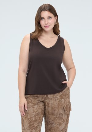 Vrouw met kastanjebruin haar tot op de schouders, die een donker mouwloos topje en een bruine broek met bloemenpatroon draagt, staand met een hand in de zak.