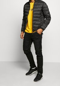 Schwarze Steppjacke mit Reißverschluss und horizontalen Nähten. Gelbes T-Shirt darunter mit schwarzer Grafik. Schwarze Cargo-Hosen mit Taschen. Schwarze Sneakers.