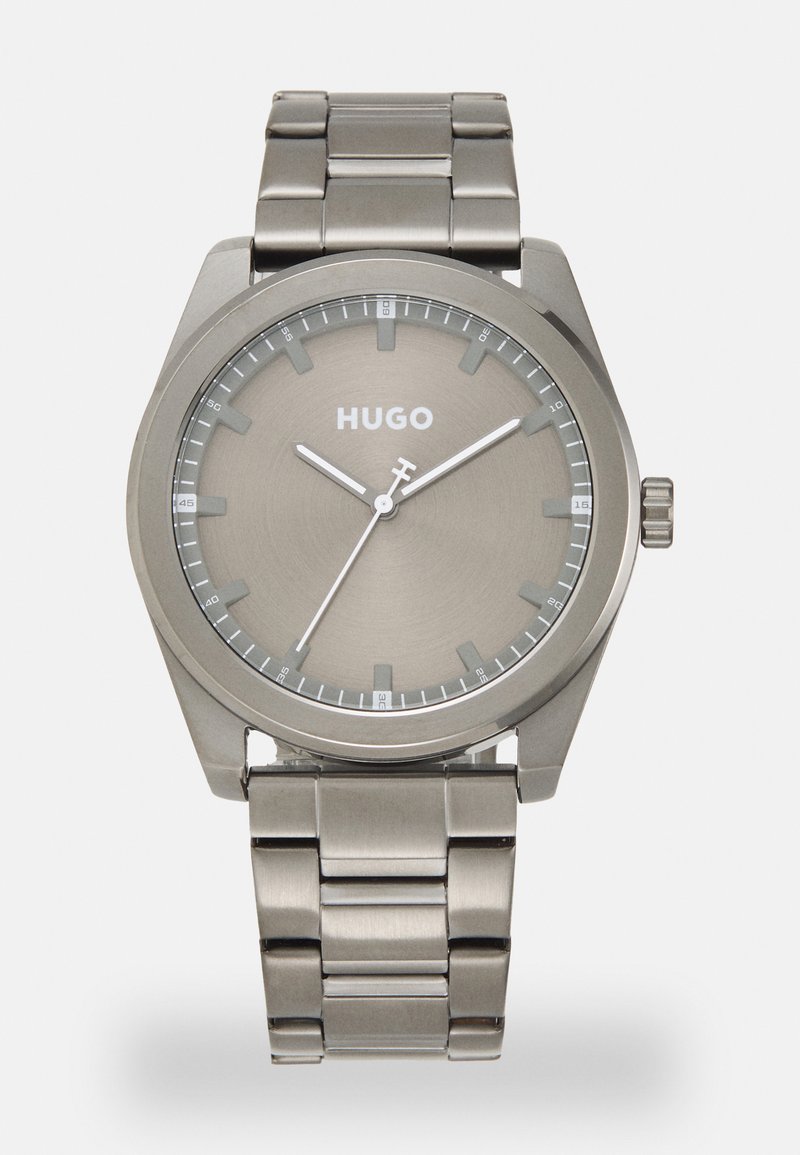 Montre en acier inoxydable argenté avec un cadran rond, un fond gris, des index blancs et un bracelet à maillons assorti. Nom de la marque "HUGO" affiché.