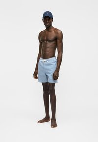 Vyriškas modelis, dėvintis mėlynai baltus languotus maudymosi šortus su balta raišteliu ir tamsiai mėlynę kepurę, basas stovintis priešais paprastą foną.