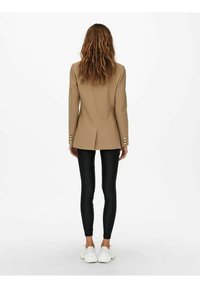 Brązowa dopasowana marynarka z poduszkami na ramionach, pojedynczym rozcięciem z tyłu i złotymi guzikami, zestawiona z czarnymi legginsami i białymi trampkami.