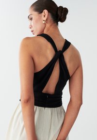 Must halterkaelusega topil on keerutatud seljaosa disain ja tekstureeritud kangas. Modell kannab helekat seelikut, mis loob tooni kontrasti.