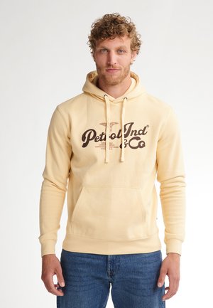 Sudadera amarilla clara con cordones, bolsillo delantero y texto gráfico en marrón. Tejido suave, mangas largas y puños acanalados en la parte inferior.