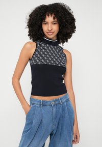 Top sin mangas de punto azul marino con un patrón geométrico texturizado y cuello alto; combinado con pantalones de mezclilla de tiro alto en azul claro.