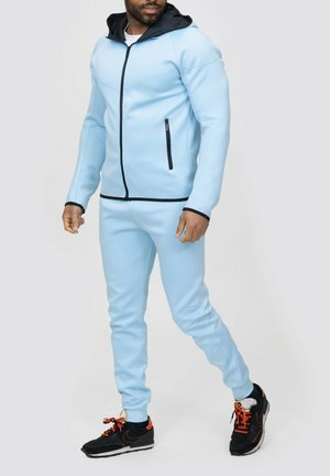 Survêtement de sport bleu clair avec accents noirs, poches zippées et capuche. Design ajusté avec une texture lisse. Chaussures de sport noires et orange.