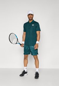 Teal atletisk polo skjorte med diamantmønster, matchende shorts, hvid kasket og sorte sneakers, mens der holdes en tennisracket.