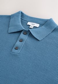 Teal ribbad pikétröja med en mjuk krage, utrustad med tre svarta knappar och en etikett med texten "NEXT", storlek M.