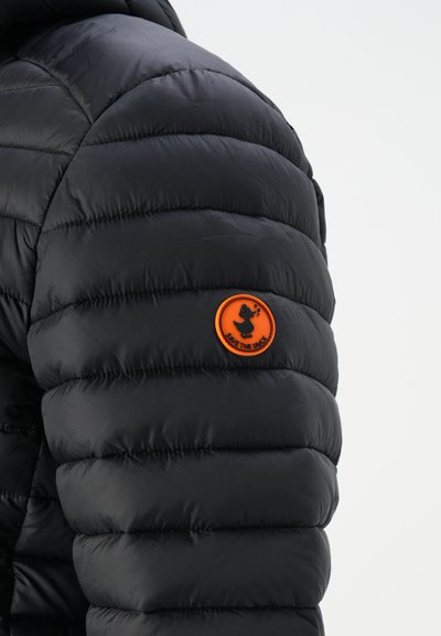 Veste rembourrée noire avec un design matelassé et une texture brillante. Présente un patch rond orange portant la mention "SAVE THE DUCK."