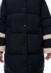 Zwarte gewatteerde jas met een boxy vorm, voorzien van een knoopsluiting en crèmekleurige faux bont accenten op de mouwen. Gestructureerde, geïsoleerde stof.