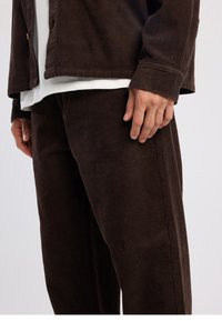 Ensemble chemise et pantalon en velours côtelé marron, présentant une texture côtelée verticale, une coupe décontractée et une couche intérieure blanche visible à l'ourlet.