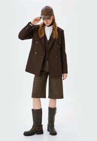 Blazer marrone a doppio petto sopra un gilet scuro e una camicia bianca, abbinato a pantaloni corti a quadri marroni, stivaletti in pelle marroni e un cappellino.
