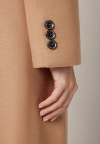 Rękaw płaszcza z wełny wielbłądziej z trzema ciemnymi guzikami, gładka tekstura i dopasowany krój w pobliżu mankietu.