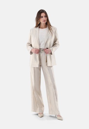 Frau trägt beigen, strukturierten Anzug mit weitem Hosenbein und übergroßem Blazer, kombiniert mit weißem, gemustertem Oberteil und beigen spitzen Absätzen.