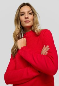 Roter Pullover mit Reißverschluss, strukturierter Oberfläche, hohem Kragen und langen Ärmeln. Verfügt über eine Tasche und ein kleines Emblem auf dem Ärmel.