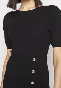 Femme portant une robe noir côtelée à manches courtes avec épaules bouffantes et trois boutons argentés sur le panneau de la taille.