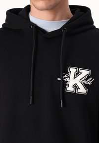 Sweatshirt negru cu glugă, având un logo brodat alb "K", șnururi ajustabile și un finisaj de țesătură moale și texturată.