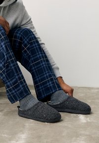 Sivi filcasti copati z gumijastim podplatom, zaobljenim prstom in subtilnimi detajli šivov. Nošeni preko modrih kariranih pižamnih hlač in sivih nogavic.