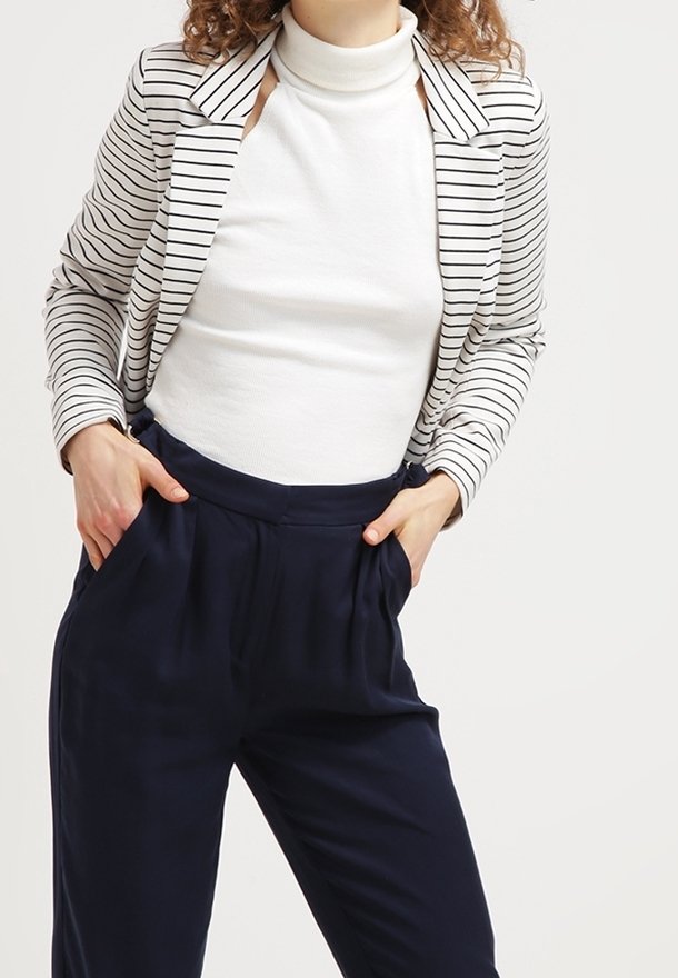 Femme portant un col roulé sans manches blanc, un blazer rayé blanc et noir, et un pantalon taille haute bleu marine avec les mains dans les poches.