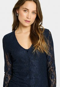 Navy lace cardigan with long sheer sleeves, V-neckline, and button-front closure. Features floral texture and a fitted design.