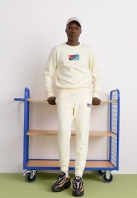 Lys beige Nike sweatshirt og matchende sweatpants, der har et lille multicolor logo, båret med sorte og guld sneakers.