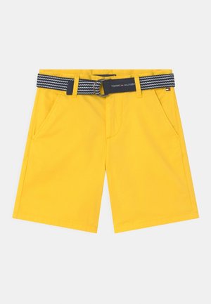 Gule shorts med en glat tekstur, der har sidelommer og et mønstret sort-hvidt bælte med en rund spænde.