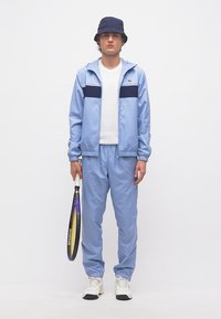 Νεαρός άνδρας με ανοιχτό μπλε φόρμα, λευκά αθλητικά παπούτσια, σκούφο τύπου κάπελο μπλε ναυτικού, κρατώντας ρακέτα τένις στο πλάι του, σε απλό λευκό φόντο.