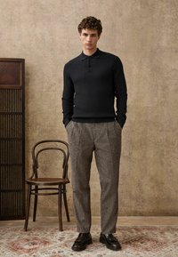 Dark ribbed long-sleeve polo shirt paired with light gray, pleated trousers, and black leather shoes. Background features a wooden chair.