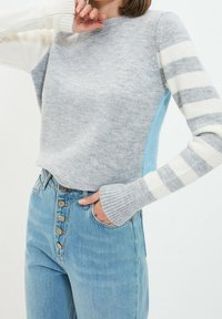 Maglione grigio lavorato a maglia con maniche lunghe a strisce bianche e grigie, caratterizzato da un pannello laterale azzurro chiaro. Abbinato a jeans blu a vita alta.