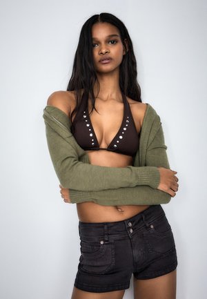 Fiatal nő hosszú hajjal, barna szegecselt bikinifelsőt, olíva zöld nyitott kardigánt és fekete farmer rövidnadrágot visel, karba tett kézzel áll.