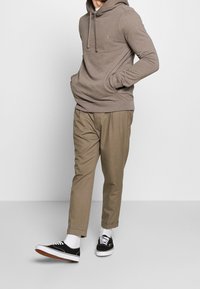 Felpa marrone chiaro con tasca a marsupio e lacci, abbinata a pantaloni beige a gamba affusolata e sneaker nere con suola bianca.