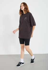 Overdimensjonert brun T-skjorte med en hvit logo på brystet, kombinert med sorte biker-shorts og sorte joggesko, båret av en modell.