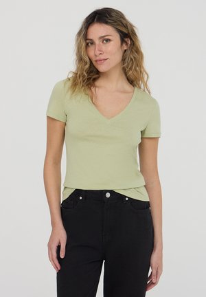 T-shirt verde chiaro a maniche corte con scollo a V realizzato in tessuto morbido, indossato con jeans neri. Presenta una vestibilità rilassata e una texture liscia.