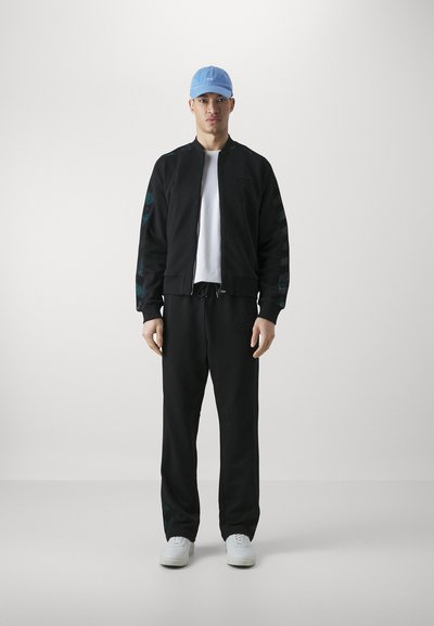 Black sports jacket with teal patterns on sleeves, white t-shirt, black sweatpants, and light blue cap, paired with white sneakers.