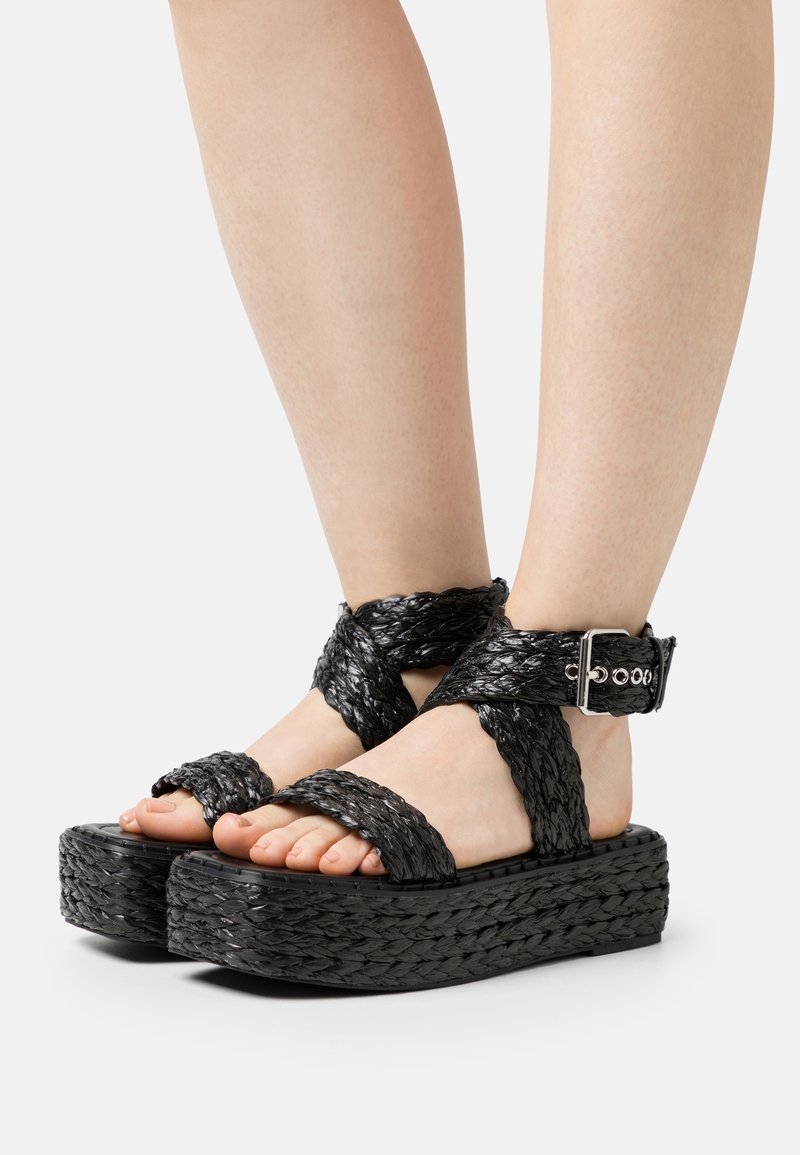 Pieds portant des sandales compensées noires tressées avec lanières à la cheville et boucle argentée sur fond clair.