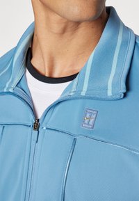 Chaqueta azul claro con cremallera, que presenta un cuello alzado, costuras horizontales y un pequeño parche con el logo de Nike. Se lleva en capas con una camiseta blanca debajo.