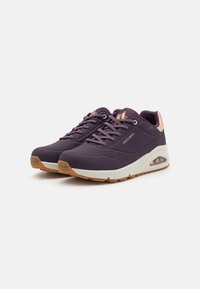 Des chaussures de sport violettes avec une finition texturée, une semelle intermédiaire blanche et des accents rose pâle. Elles présentent des détails perforés et un talon rembourré.