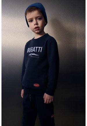 Ragazzo giovane che indossa un cappello di lana blu e felpa e pantaloni neri Bugatti, in piedi contro uno sfondo scuro uniforme.