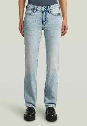 Personne portant un jean droit bleu clair et des chaussures plateformes noires, debout sur un fond uni.